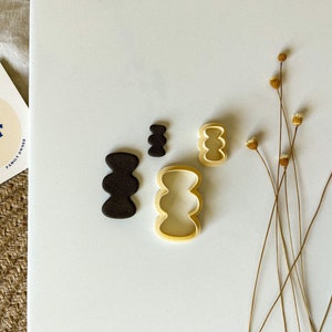 May include: A set of four cookie cutters in different sizes, featuring a wavy, organic shape. The cutters are made of gold-colored metal and are arranged on a white surface. The text "buttr cuttr FAMILY OWNED" is visible on a white card in the background.