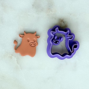 May include: A terracotta-colored cow-shaped cookie cutter and a purple cow-shaped cookie cutter. The cow has a rounded body, small horns, and a smiling face. The purple cutter has a scalloped edge.