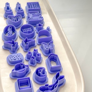 May include: A set of 17 purple plastic cookie cutters featuring various shapes, including stars, butterflies, a fish, a moon, a book, a lighthouse, a heart, a square, a planet, a crescent moon, a star, a butterfly, a bird, a ring, a star, a book, and a heart. The cookie cutters are arranged on a white surface.