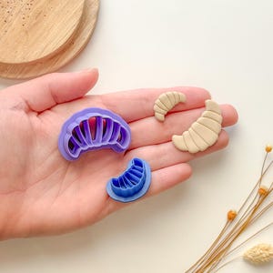 Puede incluir: Un juego de tres cortadores de galletas de plástico morado y azul con forma de croissant. Los cortadores están en una mano con un fondo marrón claro.