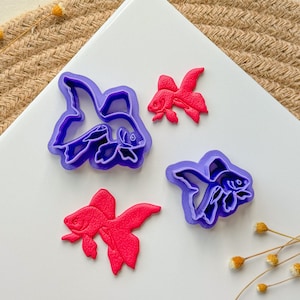 May include: Three purple and pink plastic cookie cutters shaped like goldfish. The cutters are on a white surface.