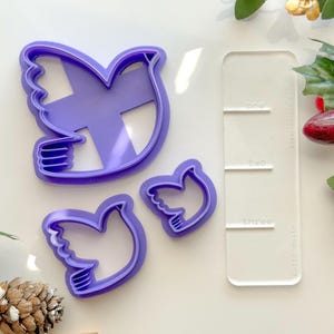 May include: Set of three purple plastic dove cookie cutters. The largest cutter is approximately 4 inches tall and 3 inches wide. The two smaller cutters are approximately 2 inches tall and 1.5 inches wide. The cutters are on a white surface with a clear plastic ruler.