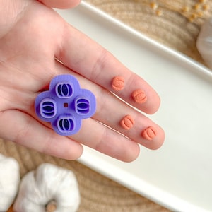 May include: Purple plastic pumpkin cookie cutter with four individual cutters. Four orange pumpkin-shaped cookies are shown next to the cutter.