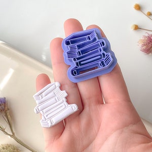 Puede incluir: Dos cortadores de galletas con forma de libro, uno morado y otro blanco. Los cortadores se sostienen en una mano, mostrando el detalle de los libros apilados.