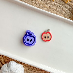 May include: Two apple-shaped cookie cutters, one purple and one red, on a white surface.