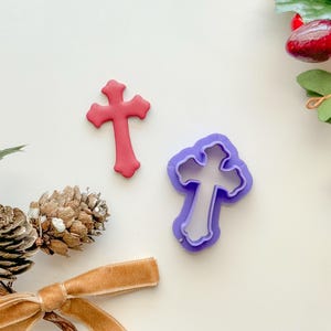 May include: A purple plastic cross cookie cutter and a red cross cookie. The cookie cutter is on a white surface with a brown ribbon and pine cones.