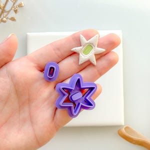 May include: A collection of polymer clay cutters in various shapes. Includes a purple star-shaped cutter, a small zero-shaped cutter, and a star-shaped embellishment with a green center. The items are displayed on a white surface.