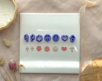 May include: A white surface with two rows of purple and pink cookie cutters. The top row includes lightning bolts, a cloud, a peace sign, a smiley face, a heart, and a mushroom. The bottom row includes lightning bolts, a flower, a peace sign, a smiley face, a heart, and a mushroom.