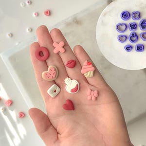 Op de afbeelding: Een hand die verschillende roze en witte polymer clay charms vasthoudt, waaronder een donut, een hart, een cupcake, een flesje toverdrank, een bloem, een lip en een kruis. De charms zijn klein en gedetailleerd, perfect om een vleugje whimsy toe te voegen aan elk project.
