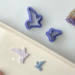 May include: Four purple and blue plastic cookie cutters shaped like birds. The cutters are on a white surface.