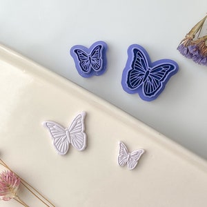May include: Two purple plastic butterfly cookie cutters and one white plastic butterfly cookie cutter on a white surface.