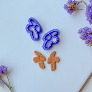 Pode incluir: Dois cortadores de biscoitos de plástico roxo em forma de flores e duas formas de flores de argila marrom.