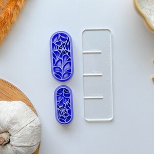 May include: Two purple plastic cookie cutters shaped like a spider web. The cutters are on a white surface next to a clear plastic ruler with the words "one", "two", and "three" printed on it.