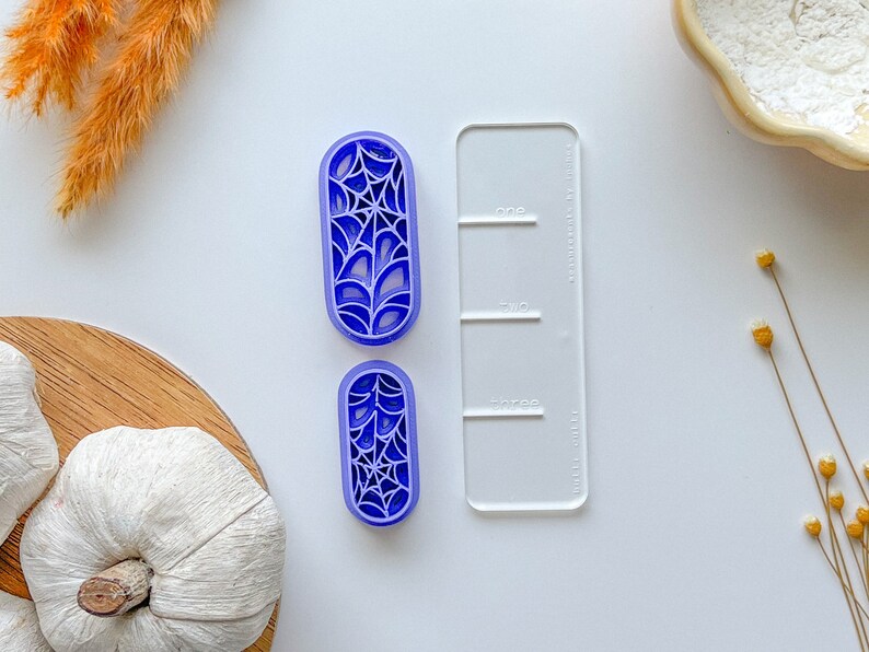 May include: Two purple plastic cookie cutters shaped like a spider web. The cutters are on a white surface next to a clear plastic ruler with the words "one", "two", and "three" printed on it.