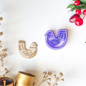 Puede incluir: Un cortador de galletas de plástico morado y un adorno de pájaro de cerámica blanca, ambos con forma de pájaro con un diseño estilizado. El adorno tiene detalles dorados.