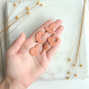 May include: A collection of small, terracotta-colored clay charms in the shape of a witch's hat, crescent moon, potion bottle, star wand, and a cauldron. The charms are displayed on a white marble surface, with dried flowers in the background.