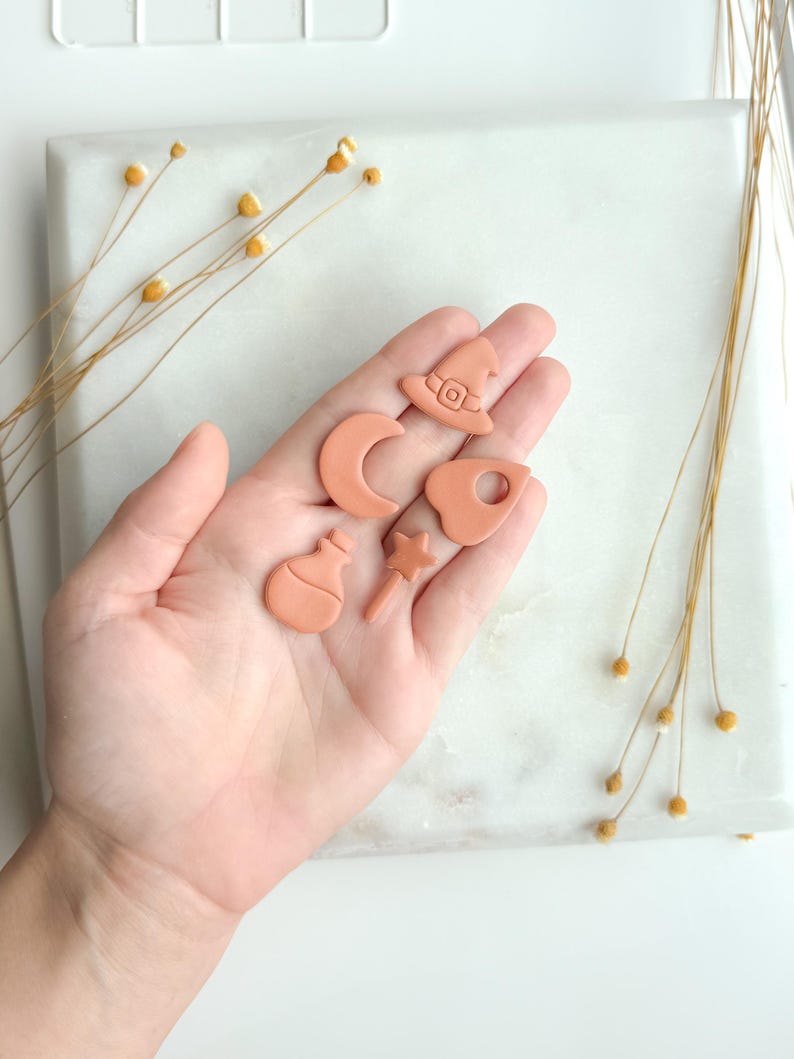 May include: A collection of small, terracotta-colored clay charms in the shape of a witch's hat, crescent moon, potion bottle, star wand, and a cauldron. The charms are displayed on a white marble surface, with dried flowers in the background.