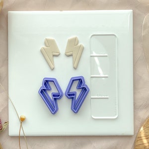 May include: Two purple plastic lightning bolt cookie cutters and two beige clay lightning bolt shapes on a white surface with a ruler.
