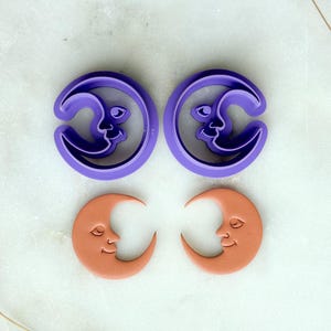 May include: Two purple moon-shaped cookie cutters and two terracotta-colored moon-shaped clay pieces. The cookie cutters have a crescent moon design with a face. The clay pieces have a crescent moon design with a smiling face.