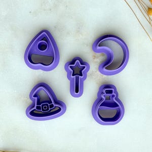 May include: Five purple cookie cutters in various shapes: a witch hat, a crescent moon, a potion bottle, a star-topped wand, and a planchette. The cutters are arranged on a white surface, with dried flowers in the background.