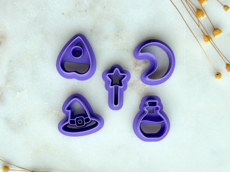 May include: Five purple cookie cutters in various shapes: a witch hat, a crescent moon, a potion bottle, a star-topped wand, and a planchette. The cutters are arranged on a white surface, with dried flowers in the background.