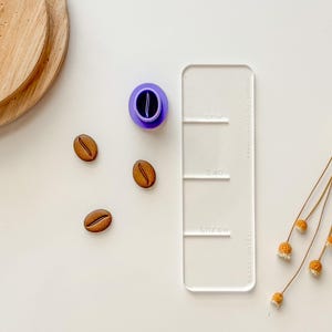 May include: A white plastic ruler with three sections labeled "one", "two", and "three". There are four brown coffee bean shaped objects on a white surface. A purple ink pad is also on the surface. A dried flower arrangement is in the background.