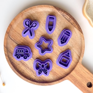 Puede incluir: Un conjunto de cortadores de galletas de plástico morado en varias formas, incluyendo tijeras, un lápiz, una estrella, un autobús escolar, un lazo, un bote de pegamento y un lápiz. Los cortadores están en una bandeja de madera.