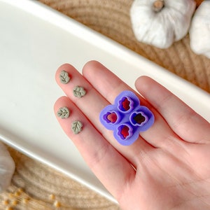 Puede incluir: Molde de plástico morado con cuatro recortes en forma de corazón. Cuatro piezas de arcilla en forma de hoja gris están sobre una superficie blanca.