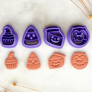 May include: A selection of Halloween cookie cutters in purple and orange. The cutters feature designs such as a witch's hat cupcake, candy corn, a jack-o'-lantern pie slice, and a bat. The orange shapes are the cut-out cookies.
