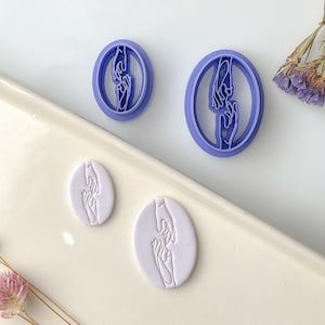 Könnte beinhalten: Zwei lilafarbene Plastik-Ausstecher in Form von Händen, die sich gegenseitig erreichen. Zwei weiße Fondant-Kekse mit dem gleichen Design sind auf einer weißen Oberfläche zu sehen.