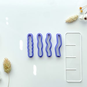 May include: Four purple plastic cookie cutters in different wavy designs. The cutters are arranged in a row on a white surface. A white plastic ruler with black markings is to the right of the cutters.