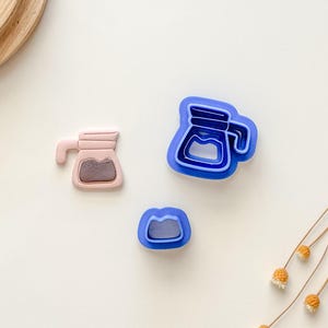Puede incluir: Tres cortadores de galletas de plástico azul en forma de cafetera, cafetera con tapa y grano de café. Los cortadores de galletas de cafetera están superpuestos.