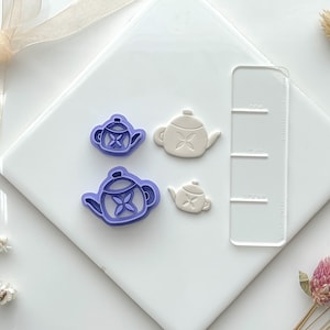 Peut inclure: Quatre emporte-pièces en plastique violet en forme de théières sur une surface blanche. Deux des emporte-pièces sont plus petits et deux sont plus grands. Les emporte-pièces sont à côté d'une règle en plastique transparente avec des marques.