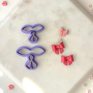 Puede incluir: Dos cortadores de galletas de lazo de plástico morado y tres lazos de fondant rosa y rojo sobre una superficie de mármol blanco.