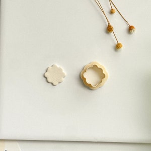 May include: A white fondant flower cutout with a plastic flower cookie cutter next to it on a white surface.