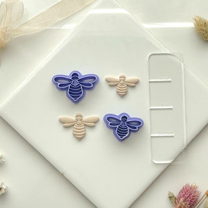 May include: Four bee-shaped cookie cutters, two purple and two beige, on a white surface. The cutters are arranged in a row with a clear plastic ruler behind them. The ruler has the words "one", "two", and "three" printed on it.