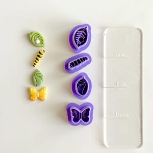 May include: An overhead shot displays a collection of colorful polymer clay cutters and finished clay pieces. The cutters are purple, shaped like leaves, a caterpillar, and a butterfly. The finished pieces are green, yellow, and white. A clear acrylic ruler is also present.