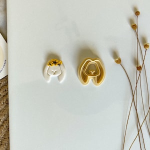 May include: Two cookie cutters in the shape of a bunny rabbit. One cutter is white with a yellow flower crown and the other is beige. Both cutters are on a white surface.