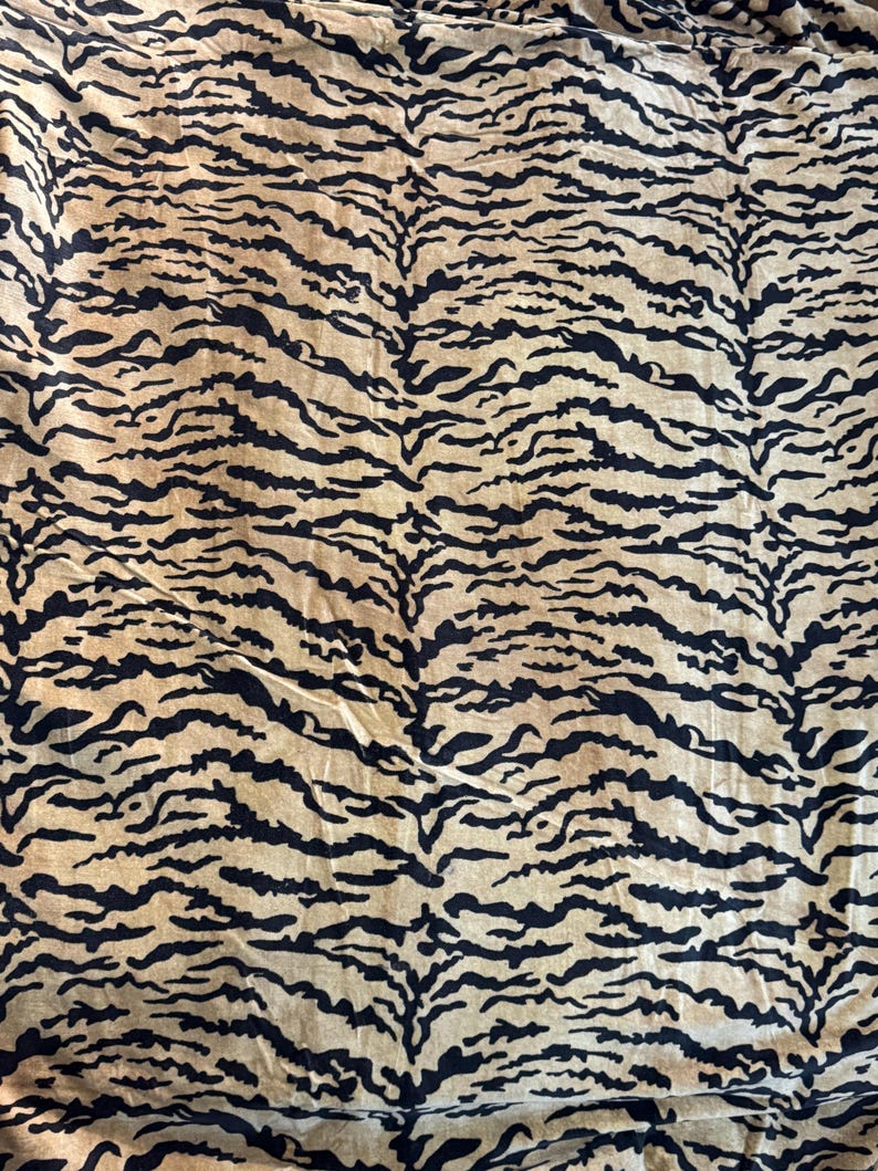 May include: A textile with a tiger stripe pattern. The fabric features black stripes on a tan background. The stripes are irregular and mimic the natural markings of a tiger. The fabric appears to be soft and pliable.