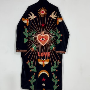 May include: A black long-sleeve coat featuring colorful embroidery on the back. The design includes a central heart with an eye, the word LOVE, a sun, birds, moths, cacti, and a crescent moon. The embroidery uses red, orange, green, and white thread.