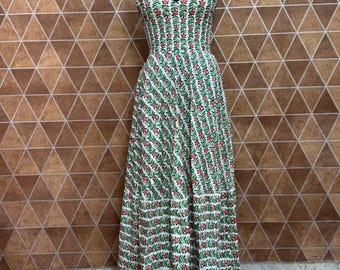 May include: A white maxi dress with a V-neck and spaghetti straps. The dress features a repeating pattern of green leaves and red flowers. The dress is displayed on a black mannequin.