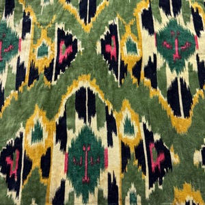 May include: A close-up of a green fabric with a detailed pattern. The design includes repeating geometric shapes in black, yellow, pink, and cream. The fabric has a soft, velvety texture, possibly for upholstery or decorative use.