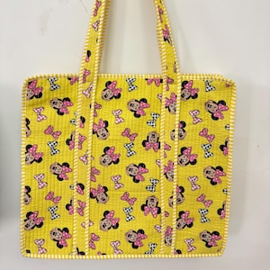 Puede incluir: Bolso tote amarillo con un patrón repetido de caras de Minnie Mouse y lazos rosas. El bolso tiene asas y ribetes a rayas amarillas y blancas. El bolso tiene forma rectangular.