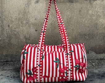 Puede incluir: Un bolso tote a rayas rojas y blancas con ilustraciones de cerezas. El bolso tiene forma rectangular, dos asas y un bolsillo delantero. El diseño de cerezas incluye cerezas rojas y hojas verdes.