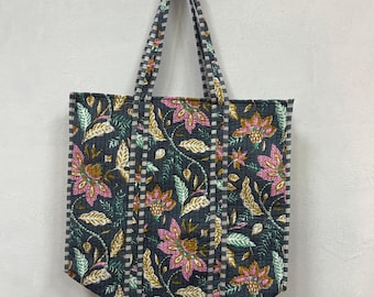 Op de afbeelding: Een gewatteerde draagtas met een bloemenpatroon in roze, goud en groen op een donkergrijze achtergrond. De tas heeft een streepjespatroon op de zijkanten en handvatten. De tas hangt tegen een witte muur.