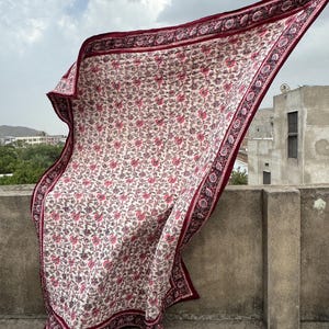 May include: A rectangular, cream-colored textile with a floral pattern in shades of pink and purple. The border is a deep burgundy with a repeating geometric design. The fabric is billowing in the wind against a cloudy sky.