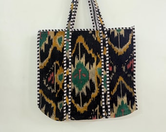 May include: A large tote bag with a black background and a colorful ikat pattern in shades of gold, green, and red. The bag has black and white striped handles and trim. The bag is made of a soft, textured fabric.