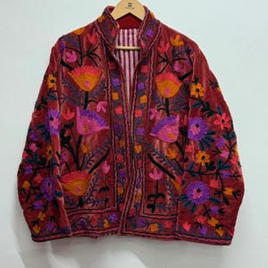 Puede incluir: Chaqueta de terciopelo burdeos con vibrantes bordados florales en tonos naranja, rosa y morado. La chaqueta presenta un cuello mandarín y un forro a rayas, creando una estética bohemia.