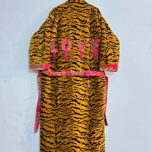 May include: A yellow and black tiger-striped robe with the word "LOVE" embroidered in pink on the back. The robe has pink trim and a matching belt. The robe is hanging on a white wall.