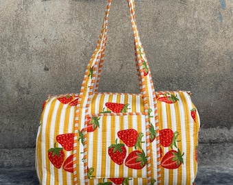Puede incluir: Un bolso tote rectangular con un estampado de rayas blancas y amarillas, adornado con ilustraciones de fresas rojas. El bolso tiene un bolsillo frontal y dos asas largas. El bolso es de tela.